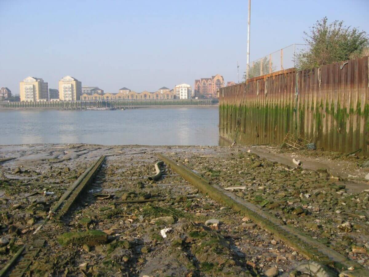 A muddy river shore, with rusty rail tracks leading into the water